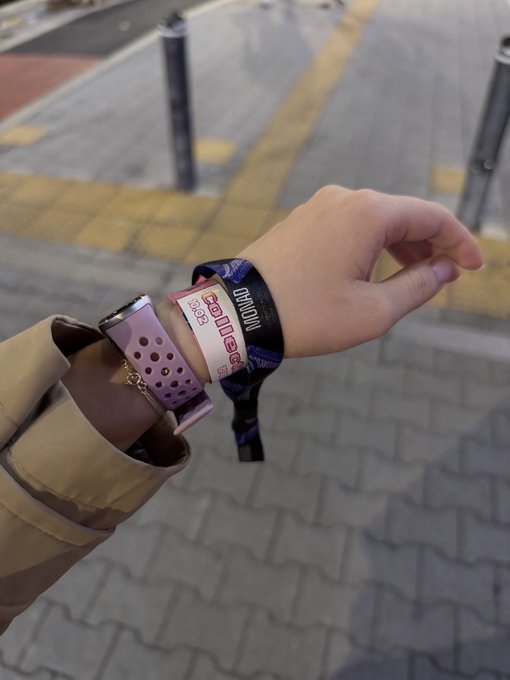 A person's arm extended outdoors wearing multiple wristbands including a black one that reads "Dominion" and pink/purple bands, with a beige bag strap visible.