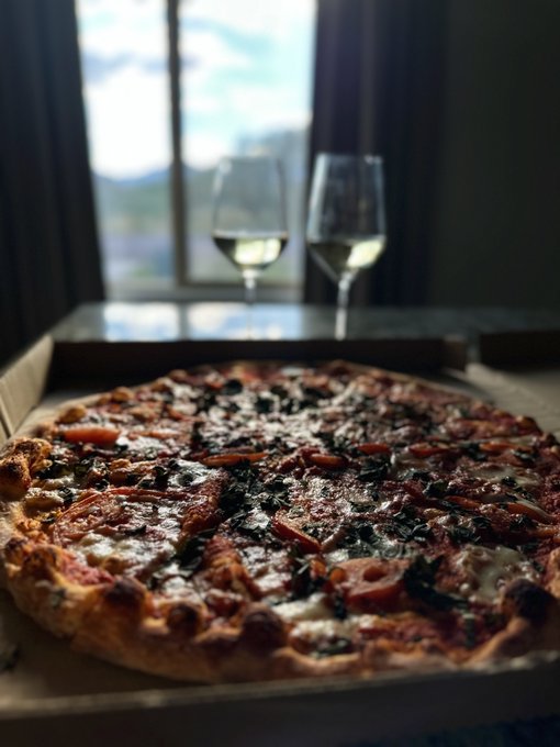 A close-up of a freshly baked pizza with toppings in an open box, set on a table with two wine glasses visible in the background near a window.