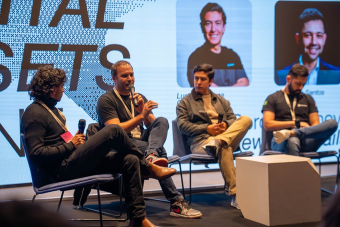 Four men sit on a stage during a panel discussion, with one speaking into a microphone, set against a large digital screen and portraits of two other men.