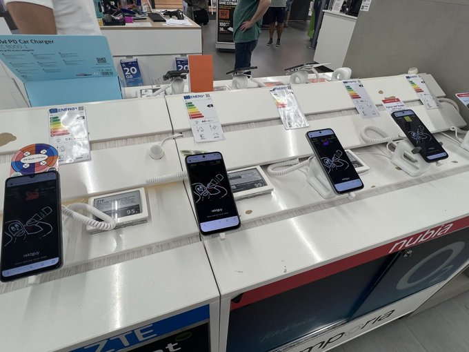 A retail store display counter showcasing several smartphones on security tethers with demo screens, accompanied by energy efficiency labels and pricing tags.