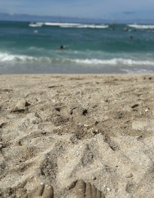 A close-up of footprints in the sand on a beach, with the ocean waves and a swimmer blurred in the background under a blue sky.