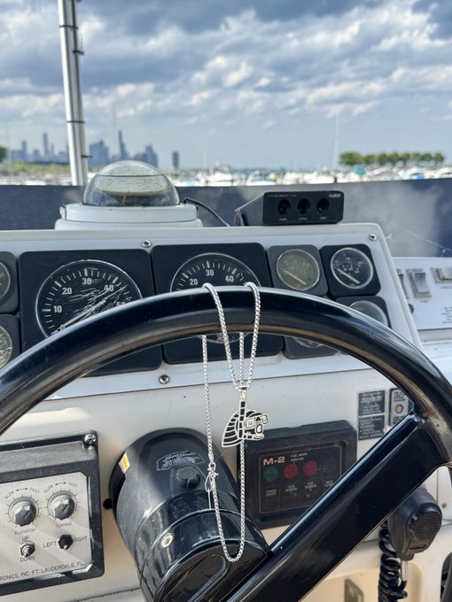 A view from the helm of a boat shows the steering wheel, instrument panel, and a Chicago Bears pendant necklace hanging from the controls, with a city skyline in the distance.
