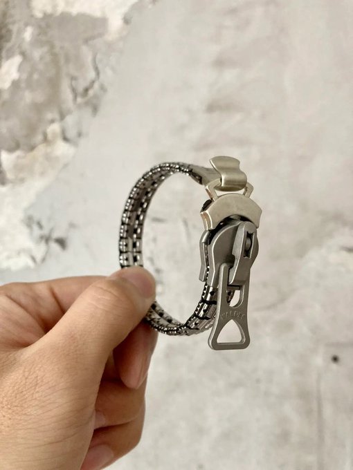 A hand holding up a silver metallic watch bracelet with an intricate link design and a distinctive deployant clasp engraved with the 'TUDOR' logo, photographed against a textured white/grey surface.