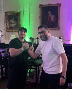 Two smiling men shake hands with a clenched fist gesture in an ornately decorated room featuring classic portrait paintings and colorful green and purple lighting, symbolizing a successful collaboration. The image commemorates the partnership behind porting Solana Playgrounds to the Seeker device, enabling developers to write, build, and deploy Solana apps directly from their phones.