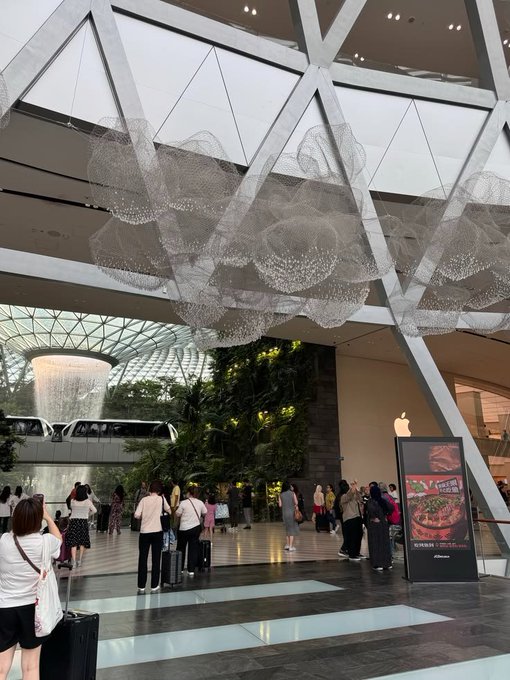 The interior of a modern shopping mall, likely Jewel Changi, featuring a striking cloud-like chandelier sculpture made of metallic mesh hanging from the glass ceiling, with an Apple Store and shoppers visible below.