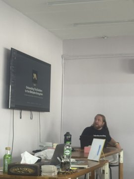 A man with long hair and beard sitting at a table in a room, with a TV screen displaying text on the wall behind him.