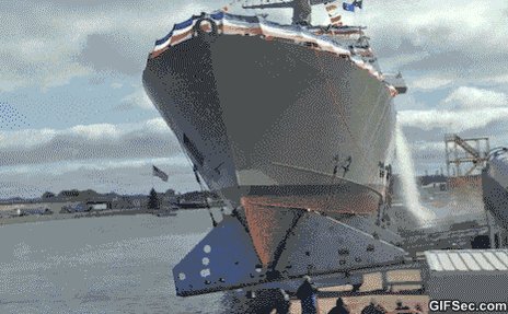 The image shows a large ship floating on water near a dock. The ship appears to be in the process of being launched or lowered into the water, possibly for an event or celebration. The scene also includes several people standing around the ship, likely observing its progress or preparing for the launch.