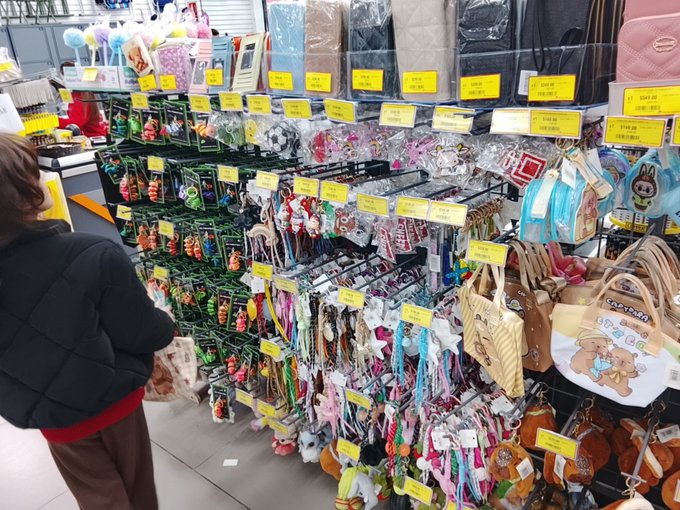 A photograph of a child in a black jacket looking at store shelves densely packed with colorful hair accessories, keychains, and small bags with yellow price tags.