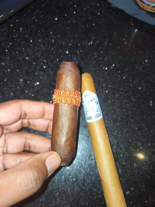 A hand holding two cigars over a black speckled countertop, one with a decorative orange and yellow band.