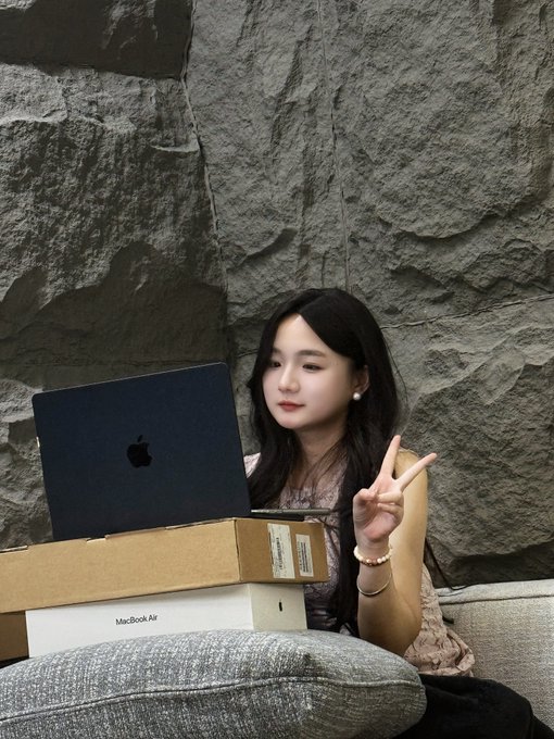 A young woman with long dark hair sits on a couch in front of a textured stone wall, smiling and making a peace sign while holding a new MacBook Air with its box.