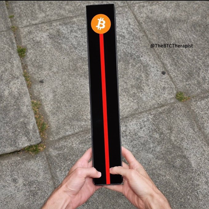 A person's hands holding a tall black box featuring the orange Bitcoin logo and a red stripe, with the watermark '@TheBCTherapist' on a stone pavement background.