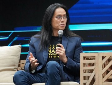 Long-haired man in glasses and a navy blazer speaking into a microphone while seated on a panel stage.