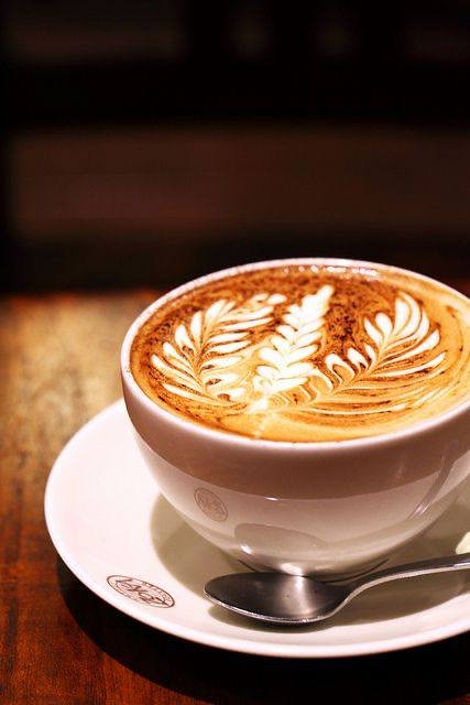 A close-up photograph of a cappuccino in a white cup and saucer on a wooden table, featuring intricate leaf-pattern latte art on the foam, with a metal spoon resting on the saucer.