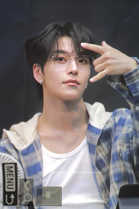 A young man with dark hair and glasses poses with his hand near his forehead, wearing a blue plaid flannel shirt over a white tee and a cross-shaped earring, while a phone screen displaying a "MENU" icon and a copyright notice ("Once in a lifetime. Forever in soul. All rights reserved.") is visible in the foreground, appearing to capture him at what seems to be an ALD1 fan event on 260829.