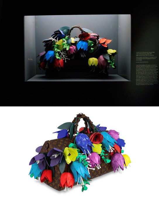 A creative Louis Vuitton x Takashi Murakami-style handbag displayed both in a museum vitrine and as a product shot, featuring the classic monogram canvas adorned with colorful 3D sculptural flowers in blue, red, yellow, purple, and green.