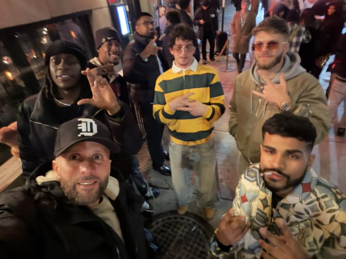 A group of six people pose together for a selfie outside at night, including a man in a Detroit Tigers cap, a man in a yellow and green striped rugby shirt, and a man in a brown Carhartt-style hoodie.