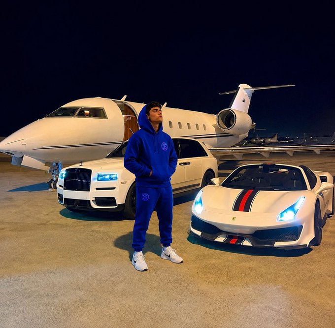 A young man in a blue tracksuit posing at night on a tarmac in front of a private jet, a white Rolls-Royce Cullinan SUV, and a white Ferrari with red and black racing stripes.