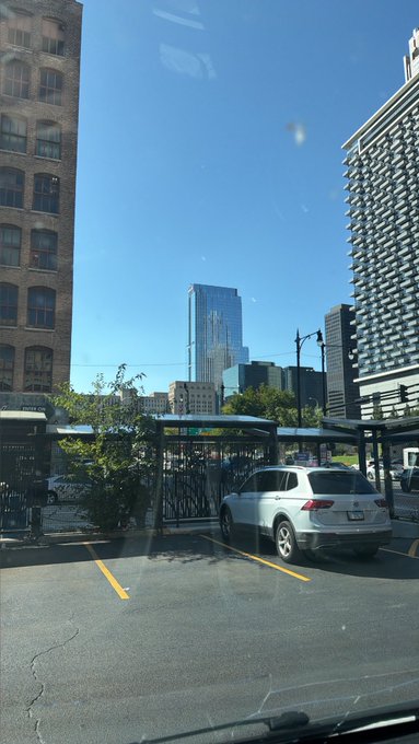 A daytime city view from inside a vehicle, looking out through a windshield at a parking lot with a white SUV, framed by tall brick and glass buildings under a clear blue sky.