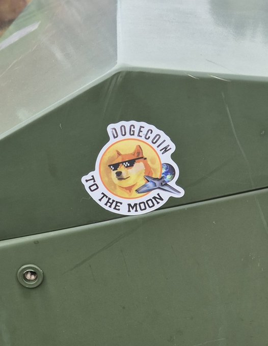 A 'DOGECOIN TO THE MOON' sticker featuring a sunglasses-wearing Shiba Inu and a rocket, placed on a green metal surface.