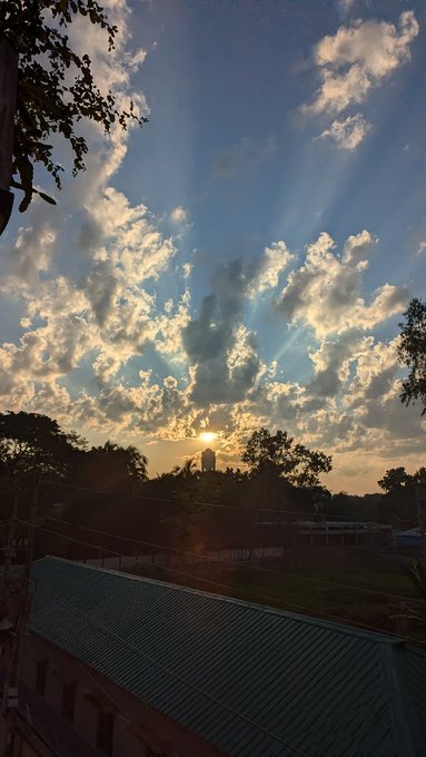 A scenic view of a sunset with rays of light shining through scattered clouds over a landscape with trees and a building roof in the foreground.