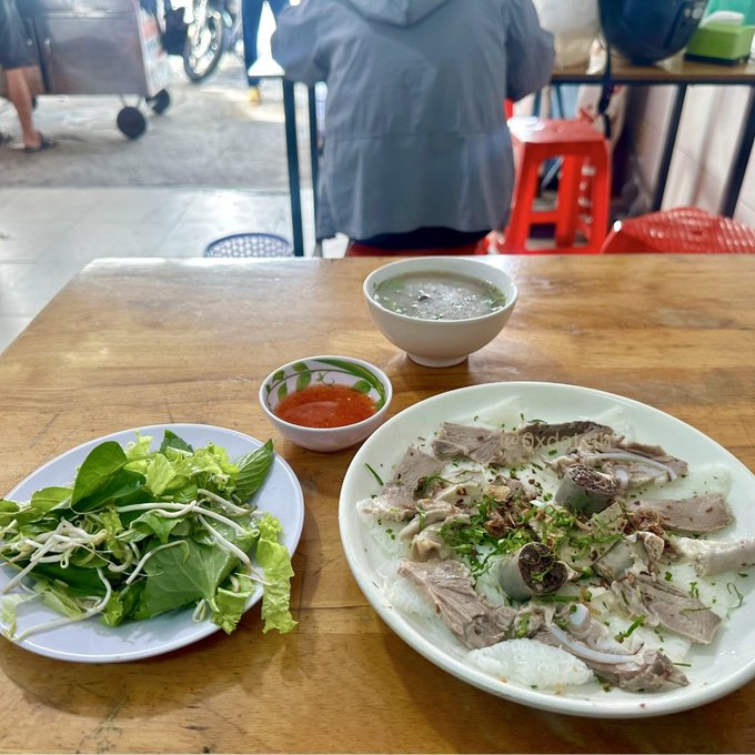 A meal consisting of a plate of sliced meat garnished with herbs, a side of fresh greens, a small bowl of red sauce, and a bowl of broth arranged on a wooden table.