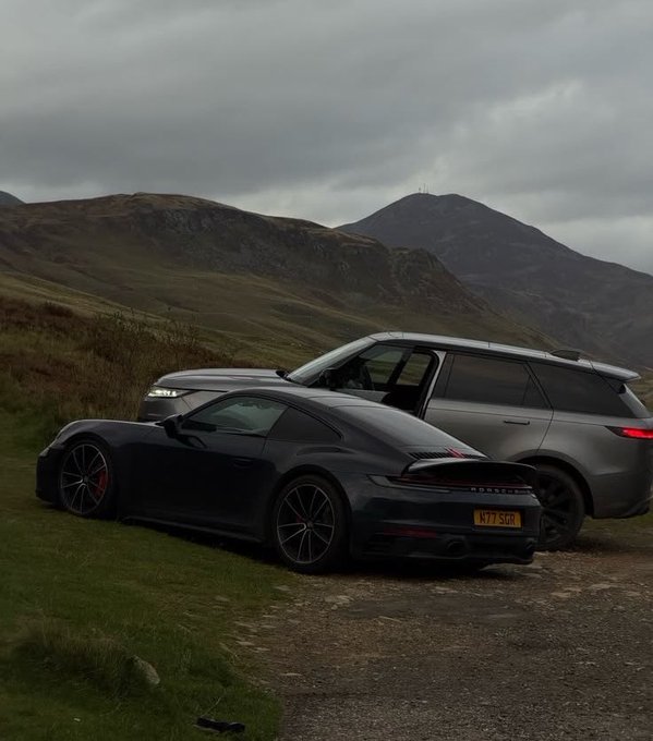 A dark gray Porsche 911 and a gray Range Rover are parked on a gravel path against a backdrop of moody, overcast mountains in what appears to be a rural setting.