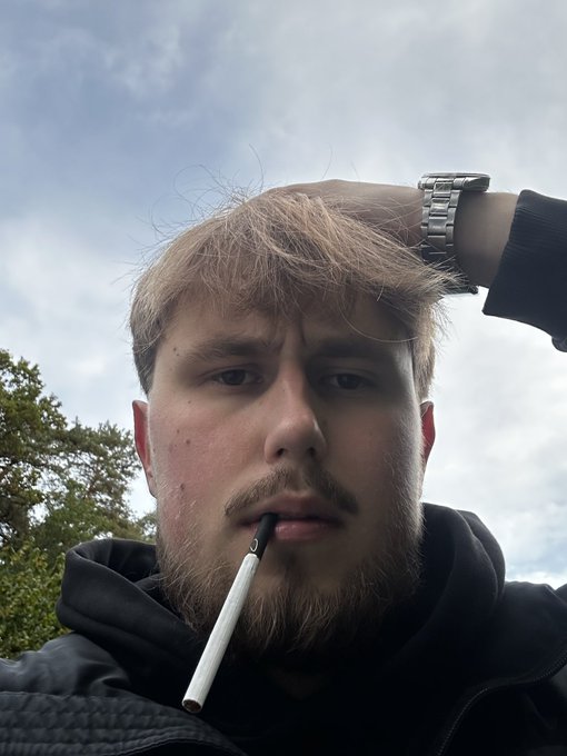 A close-up selfie of a young man with a beard and messy blonde hair smoking a cigarette, wearing a black hoodie and a metal watch, set against a cloudy sky.