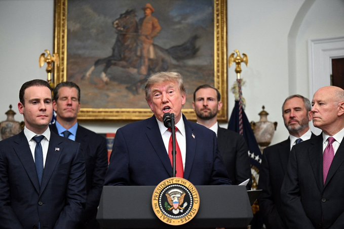 President Donald Trump speaks at a podium bearing the Seal of the President of the United States, surrounded by officials in suits, with a large painting of a cowboy on horseback hanging in the background.
