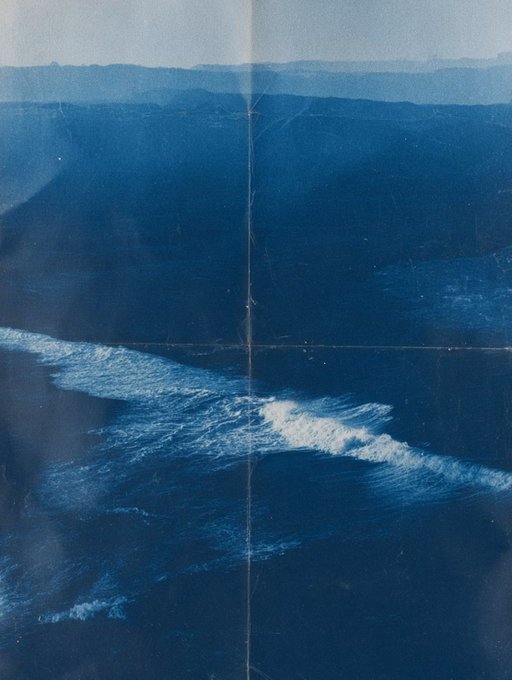 A moody blue-toned photograph or print showing an aerial view of ocean waves breaking against a dark, mountainous coastline.