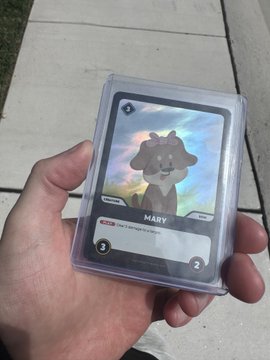 A hand holding a holographic trading card in a protective plastic case, featuring an illustration of a brown dog character named 'MARY' with a sheep subtype designation and a power level of 2.