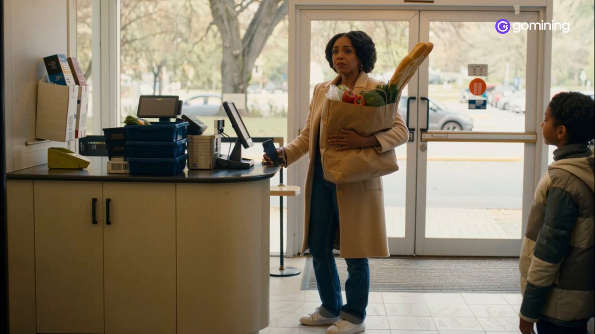 The image shows two people standing near a cash register, with one person holding a bag of groceries while the other person looks at them. The scene takes place inside a store or a market where there are various items for sale, including books and magazines on display.