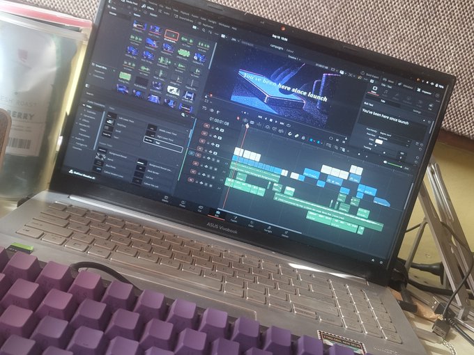 A photograph of an ASUS Vivobook laptop displaying video editing software with a timeline and preview window, accompanied by a purple mechanical keyboard and a Starbucks Roastery tumbler in the background.