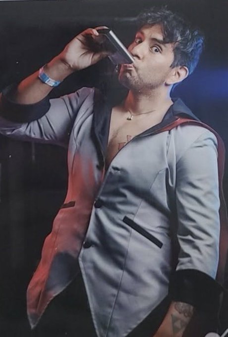 A man with dark hair in a silver satin jacket and cape drinks from a flask while making an exaggerated surprised face in a dimly lit setting.