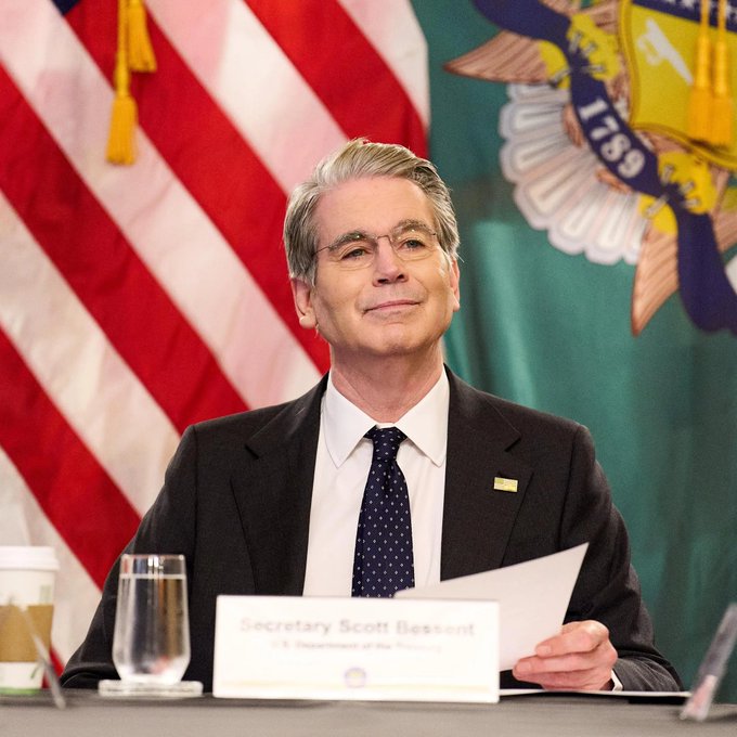 U.S. Treasury Secretary Scott Bessent sits at a table with a nameplate reading 'Secretary Scott Bessent,' holding papers, with an American flag and a Treasury Department flag displayed behind him.