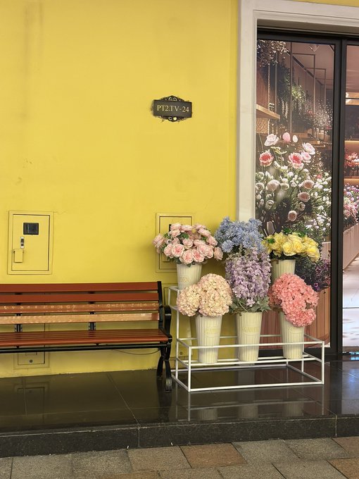 A yellow wall in what appears to be a mall corridor, featuring a wooden bench, a display of colorful flower bouquets, and a sign reading 'PT2.TV-24'.