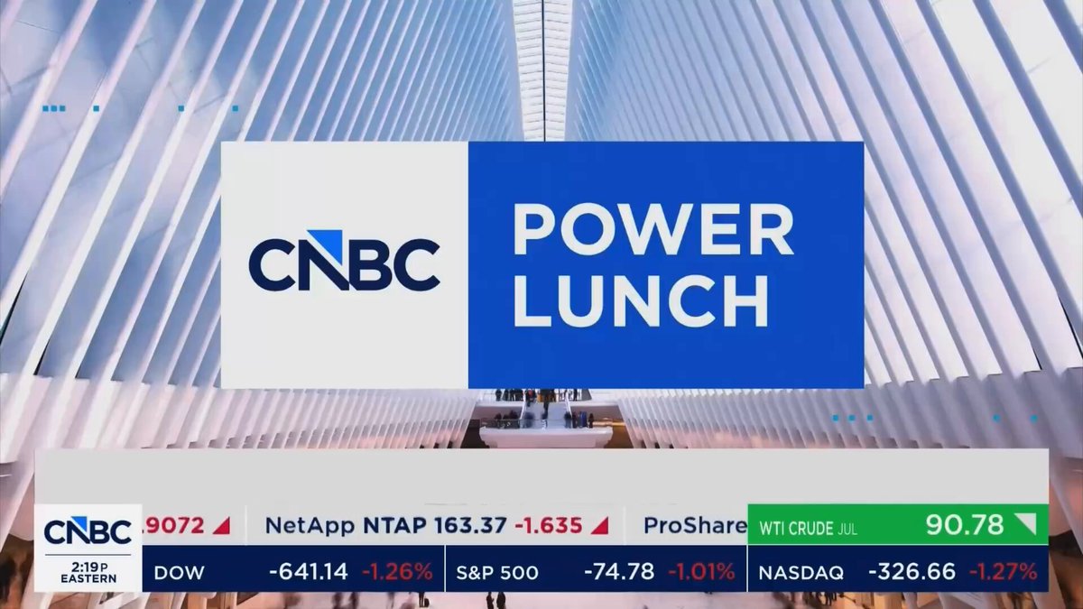 The image shows a billboard with a blue background featuring white text, including "Power Lunch" written in blue letters above a picture of a building. The text also includes mentions of Bitcoin, CNBC, and other financial terms. The image seems to be promoting an event or product related to cryptocurrency trading or investing.