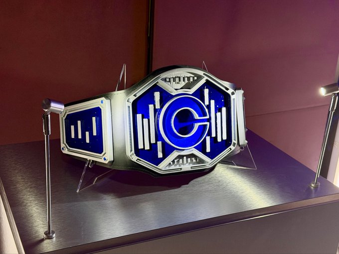 The image features a blue and silver championship belt prominently displayed on a stand, featuring a central "C" logo emblem surrounded by vertical bar designs. This appears to be the Carcosa Championship Wrestling (CCW) belt awaiting its new champion at tonight's event.
