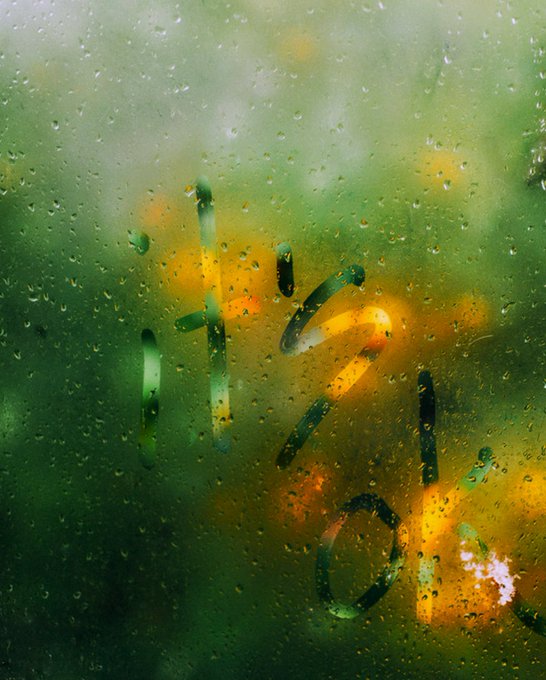 A view through a rain-covered window showing blurred green and yellow foliage outside, with 'HIGH LIFE' text visible in the background through the water droplets.
