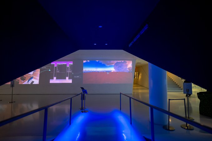 A modern museum or gallery interior features dramatic blue lighting, illuminated screens displaying digital artwork, and a staircase in the background.