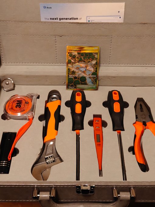 A metal toolkit case containing orange and black hand tools including a tape measure, wrench, screwdrivers, a voltage tester, and pliers, along with a Pokémon Trading Card Game blister pack featuring Charizard.