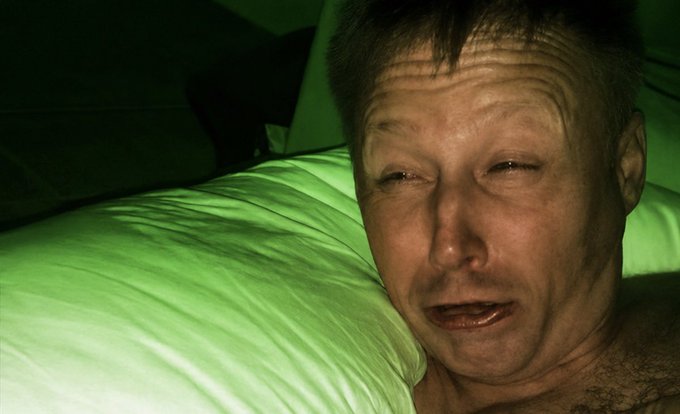 A man with a disheveled, exhausted expression lies in bed, lit by an eerie green glow, evoking the defeated and weary look of a Solana enthusiast after a tough market day.
