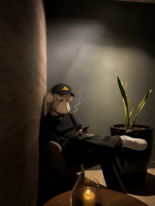 A figure wearing a Bored Ape-style mask, black clothing, and a cap sits in a dimly lit room smoking a cigarette while looking at a phone, with a snake plant and candle nearby.