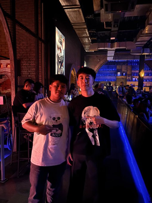 Two young men pose for a photo at what appears to be a dimly lit bar or club, with one wearing a white shirt featuring a face graphic and the other in a black t-shirt.