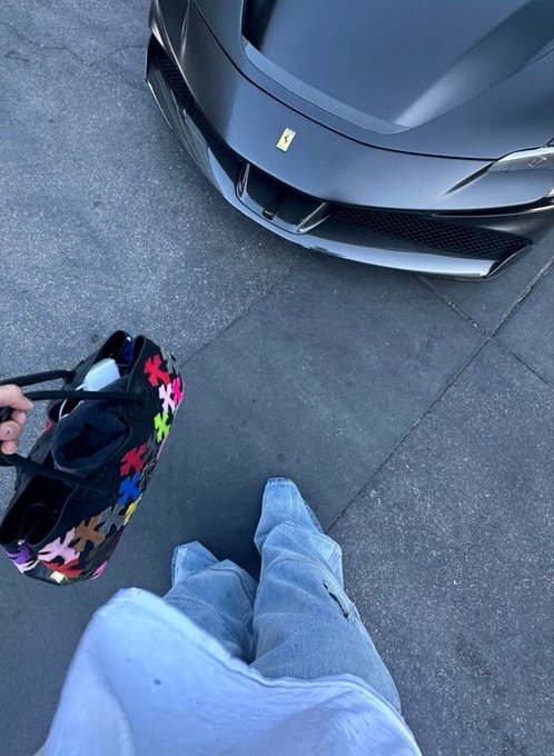 A first-person view shows someone in gray pants and holding a colorful puzzle-piece patterned bag, standing near a matte gray Ferrari sports car.