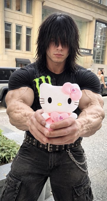 A muscular man with long dark hair wearing a black shirt holds a Hello Kitty plush toy in front of his chest on a city street.