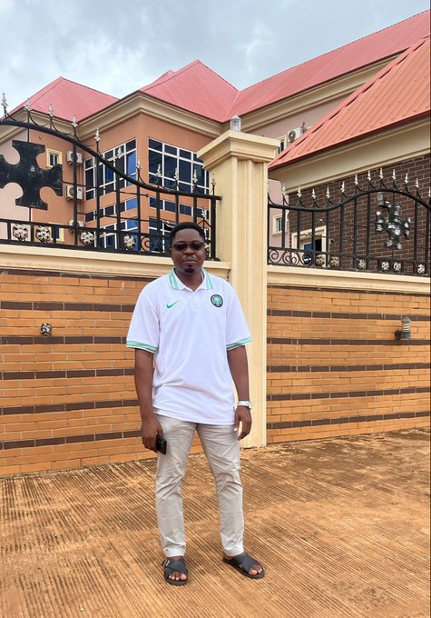 A man wearing sunglasses, a white and green polo shirt, gray pants, and sandals stands outdoors on a paved area in front of a brick wall, metal gate, and a building with a red roof.