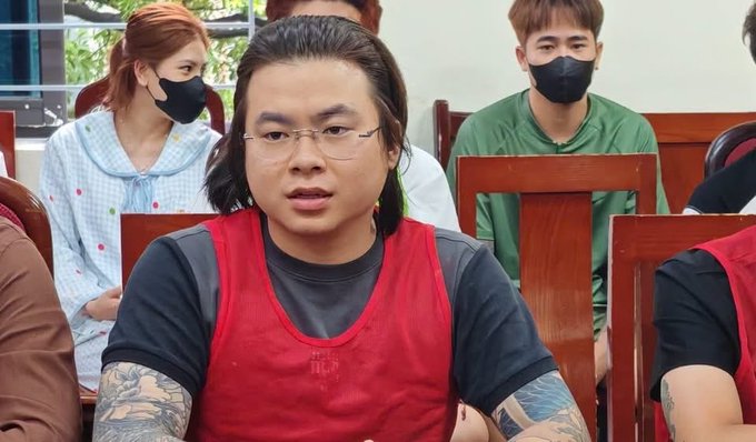 A man with glasses and a red vest sits among other people, with a person wearing a mask visible in the background.