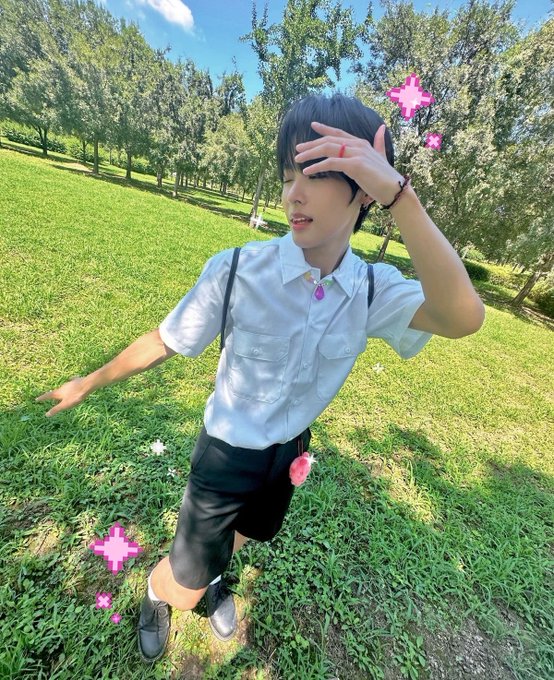 A photo of a person with short dark hair wearing a light blue short-sleeved shirt and black shorts, standing in a grassy park with trees, with pink cross-shaped filter effects overlaid on the image.