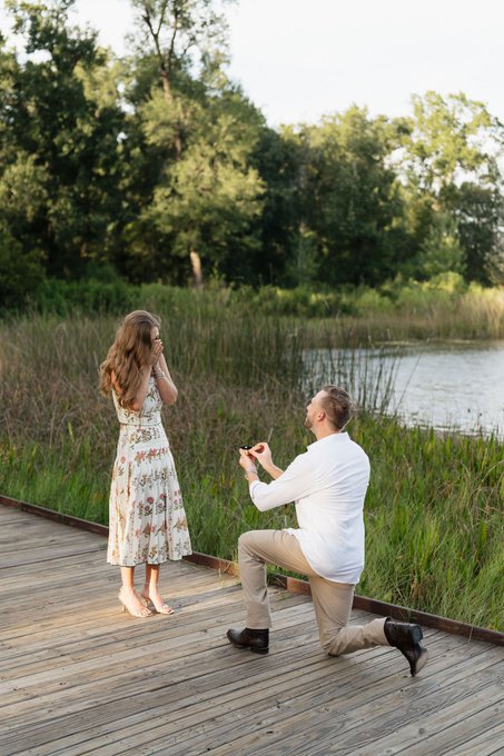 A man kneels on a wooden dock by a serene pond, presenting a ring to a woman in a floral dress who covers her mouth in surprise, capturing the moment of a proposal that declares "I win."