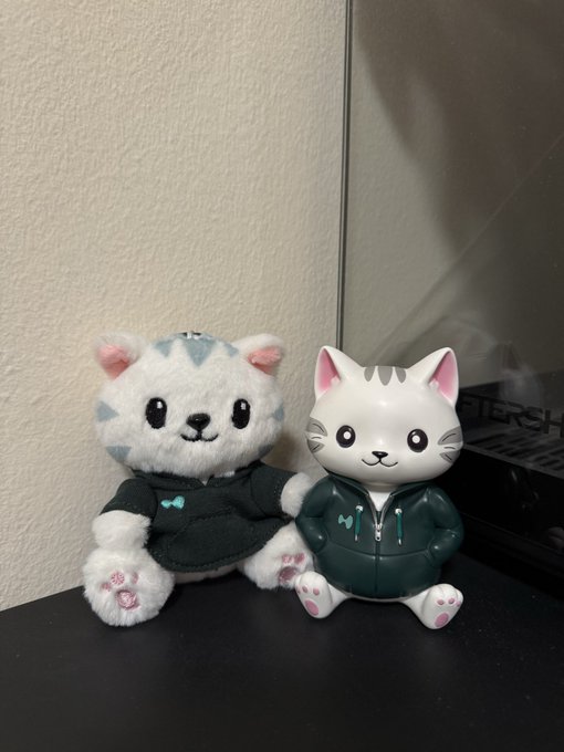 Two cat figurines sit on a desk: a plush toy in a black hoodie and a plastic cat figurine wearing a green hoodie.